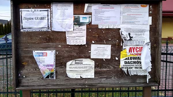 A public notice board mounted outdoors with printed sheets pinned across it