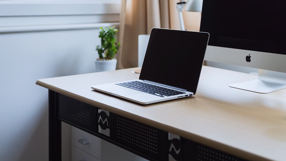 An open laptop on a tidy desk beside a notebook, lit by daylight from one side